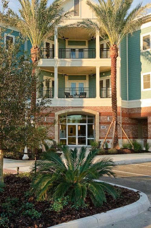 An exterior view of a 3-story apartment building at The Flats at Tioga Town Center with patios or balconies and well-maintained palm trees near resident parking.