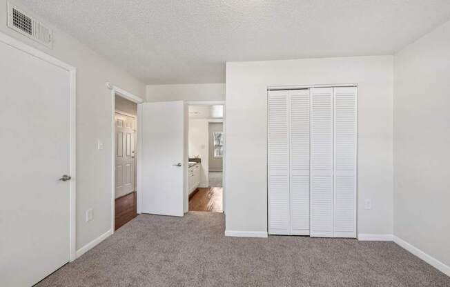 A room with white walls and carpeted floor with a white door on the left and a white closet with blinds on the right.