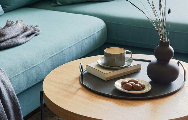 A living room with a blue couch, a wooden table with a vase, a book, a cup, and some fruit.