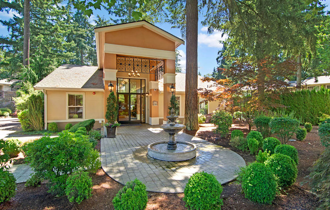 A welcoming leasing office entrance here at Asbury Park showcasing a charming building with a covered entry, glass front doors, and warm exterior accents, centered around a decorative fountain on a paved walkway, surrounded by manicured shrubs, mature trees, and landscaped greenery that create an inviting, park-like community setting.