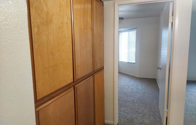 View of a hallway leading into a room with light-colored walls and carpet. On the left, there are wooden cabinets or storage units. To the right, a doorway opens into another room with a window featuring blinds, allowing natural light to enter.
