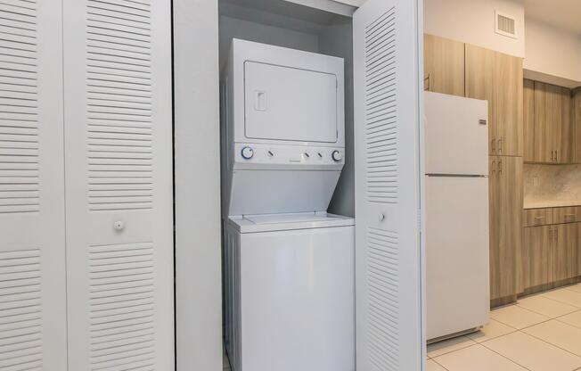 A stacked washer and dryer are located in a small closet, with doors slightly ajar. The surrounding area features light-colored walls and cabinets, and there's a refrigerator visible to the right. The floor tiles are light-colored, complementing the modern kitchen design.