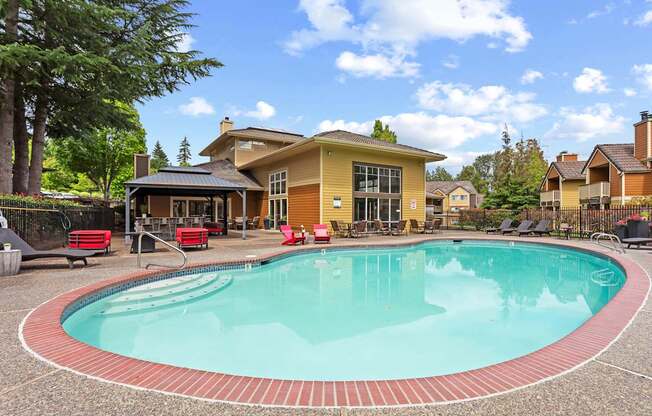A swimming pool in front of a house with a patio and lounge chairs.