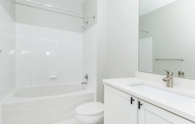 A clean, modern bathroom featuring a white tiled shower and bathtub, a white countertop with a sink and faucet, and a large mirror. The walls are painted in a light color, and there are minimalist fixtures, creating a bright and airy atmosphere.