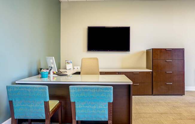 A room with a desk, chairs, and a television mounted on the wall.