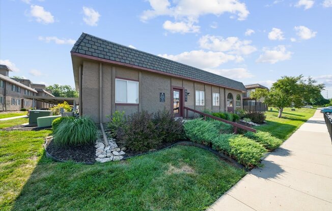 A small house with a brown roof and a red door is surrounded by a well-maintained lawn and a sidewalk.