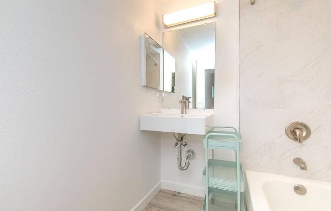 A modern bathroom featuring a sleek white sink mounted on the wall, a large mirror above, and a chrome faucet. Next to the sink, there is a light green four-tiered cart for storage. The walls are a soft white, and there is a light-colored bathtub in the corner with a simple showerhead.