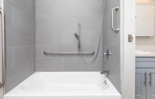 A clean bathroom featuring a modern shower-tub combo with gray tiled walls. The setup includes a handheld showerhead on a chrome bar, a grab bar for safety, and a white tub. A vanity with a mirror is partially visible in the background, showcasing a light and inviting atmosphere.