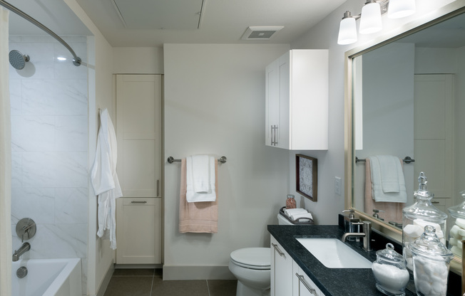 Spacious bathroom with warm gray tile floors, a large soaking tub with a shower curtain, white cabinets, black quartz countertops, and a large framed mirror.