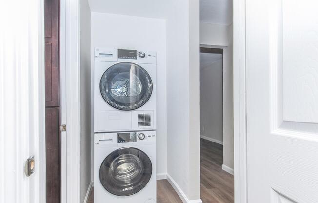 A stacked washer and dryer in a narrow hallway, with beige walls and hardwood flooring. A door is partially visible on the left, and a glimpse of another room can be seen at the end of the hallway. The space is brightly lit, creating a clean and modern laundry area.