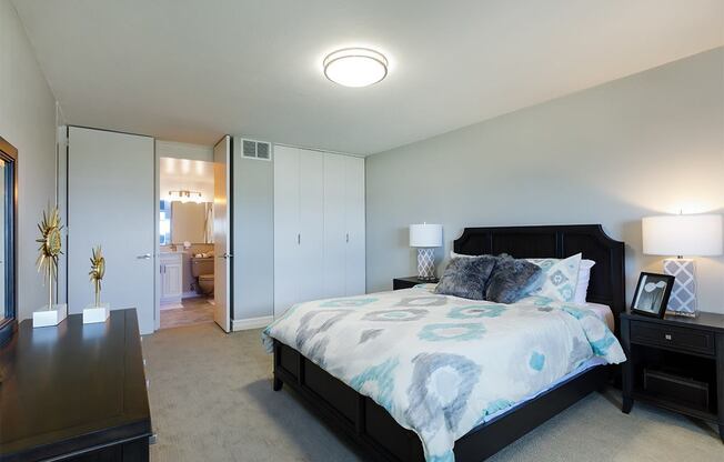 Photo of a bedroom at The Jefferson. The walls are painted a light grey. There is a queen sized bed in the middle of the room with two wood-like black end tables on each side. The bed has a patterned comforter. There is a closet with two doors that pull open. There is light brown carpet throughout the room. Next to the closet there is an enterance to the bathroom.