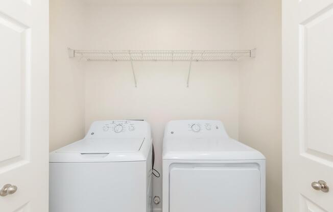 A laundry room featuring a side-by-side washer and dryer enclosed in a closet. Above them is a wire shelf for storage, and the walls are painted white. The door frames are also white, creating a clean and simple aesthetic.
