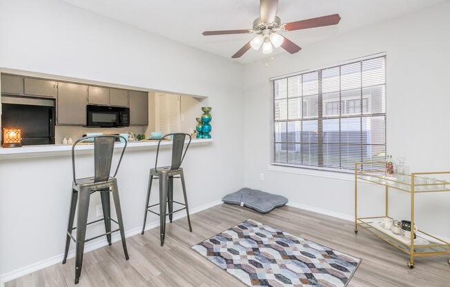 An interior view of a modern kitchen and dining area featuring two bar stools at a counter, a decorative rug on the floor, a window with blinds, and a stylish side table with decorative items. The space has neutral walls and wood-look flooring, illuminated by a ceiling fan.