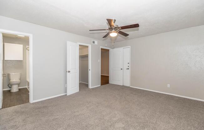A spacious, well-lit room featuring light gray walls and a ceiling fan. Two white panel doors lead to separate compartments, one likely a closet and the other a bathroom. The floor is covered in soft gray carpet, creating a cozy atmosphere.