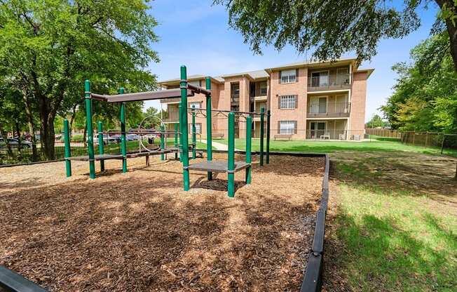A playground with a green swing set and a brown mulched area in front of a brick apartment building.