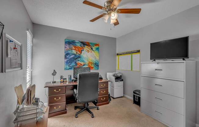 A room with a ceiling fan, a desk with a chair, a filing cabinet, and a television.