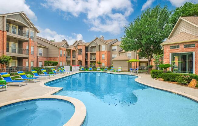 A large swimming pool surrounded by lounge chairs and umbrellas, situated in a residential area. Nearby, there are brick apartment buildings with balconies and well-maintained landscaping. The sky is partly cloudy, creating a bright, inviting atmosphere.