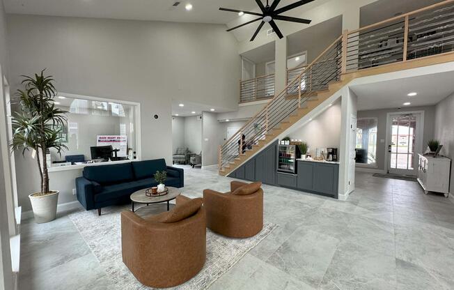 Spacious modern interior featuring a large common area with a high ceiling, a stylish staircase, two brown armchairs, a round coffee table, and a blue couch. Large windows provide natural light, and there are potted plants for decoration. A kitchenette is visible in the background.