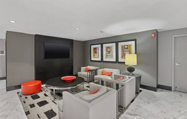 A modern living room with a grey couch, a black and white rug, a red ottoman, a black coffee table, a flat screen TV, and framed pictures on the wall.