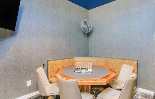 A small, modern meeting room features a circular table with a gray tabletop and wooden trim, surrounded by light-colored leather chairs. A wall-mounted fan is visible in the corner, and a flat-screen television is mounted on the opposite wall. The room has a minimalist design with gray walls.