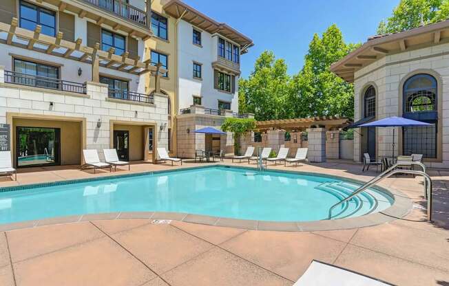 A large swimming pool is surrounded by lounge chairs and umbrellas in front of a building.