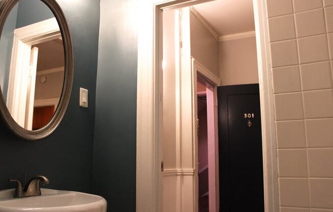 Bathroom in one bedroom brookmore apartment in Pasadena. Oval mirror over pedestal sink and doorway with white trim leads to hallway and front door.