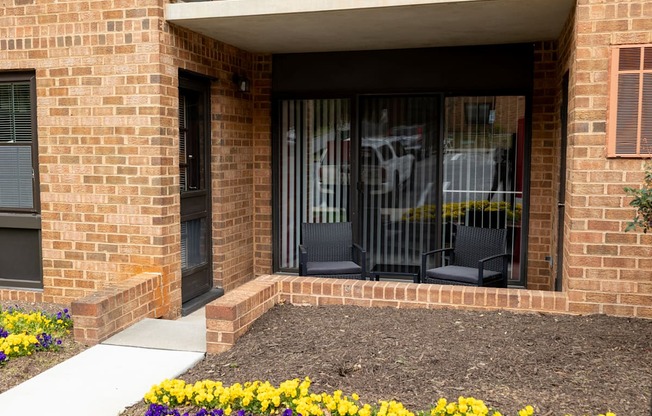 a front porch of a brick building with two chairs and flowers