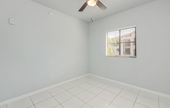 A light blue empty room with white tiled flooring, featuring a ceiling fan and a window with blinds. The walls are clean and clear of decorations, creating a minimalist and spacious feel. Natural light enters through the window, illuminating the room.
