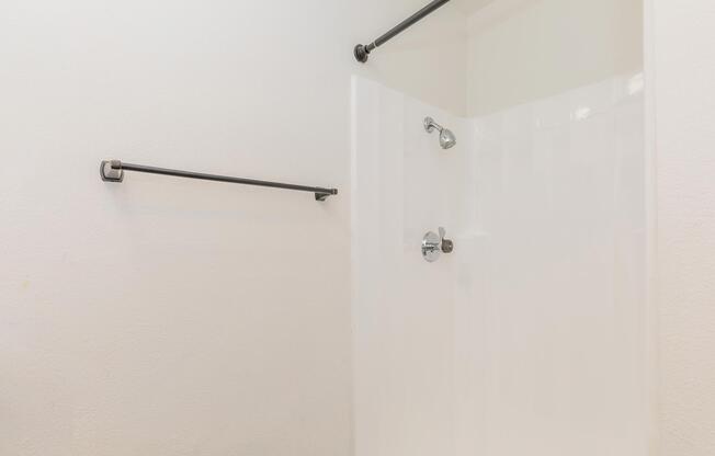 A clean, modern shower area featuring a white shower wall, a chrome showerhead, and two black towel bars mounted on the wall. The overall design is minimalistic with neutral colors, focusing on functionality and simplicity.
