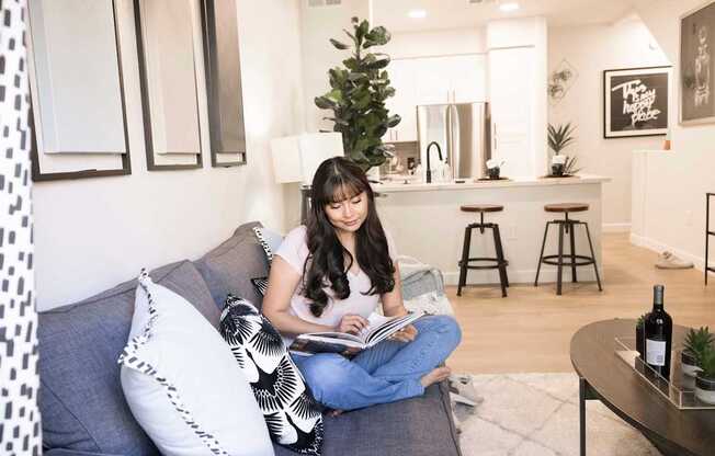 A woman is sitting on a grey couch reading a book.