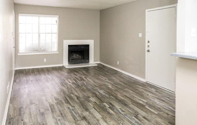 A spacious, empty living room featuring light gray walls and wooden flooring. A fireplace is centrally located on the far wall, accompanied by a window with white blinds that lets in natural light. The entrance door is visible on the right, making the space feel inviting.