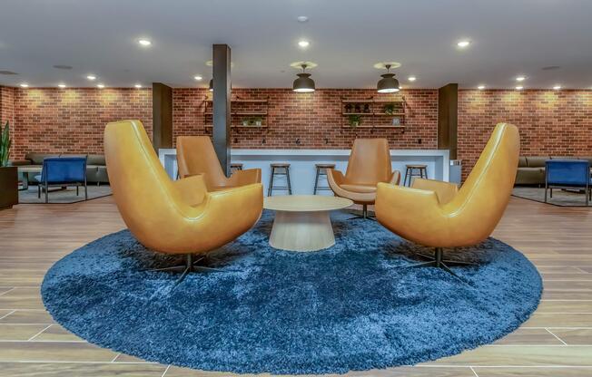 A modern lounge area featuring four stylish orange armchairs arranged around a round wooden table on a blue circular rug. The background includes a brick wall and subtle lighting fixtures, creating a cozy and inviting atmosphere. Soft seating is visible in the distance.