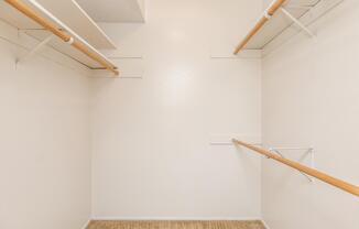 An empty closet with white walls, a beige carpeted floor, and two wooden rods for hanging clothes. There are no shelves or items visible inside, creating a spacious and minimalistic appearance.