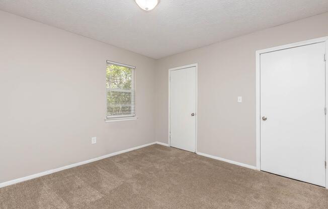 Empty room with light beige walls and a light brown carpet. One window on the left side with blinds partially drawn, and two white doors—one leading to a closet and the other is a regular door on the right. Ceiling light fixture is visible. Natural light comes in from the window.