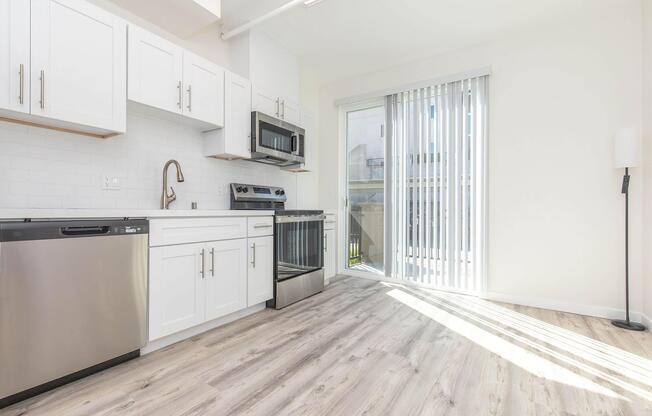 Modern kitchen featuring white cabinetry, stainless steel appliances including a microwave and dishwasher, and a sliding glass door leading to an outdoor area. The floor is light-colored wood, and the space is bright with natural light.