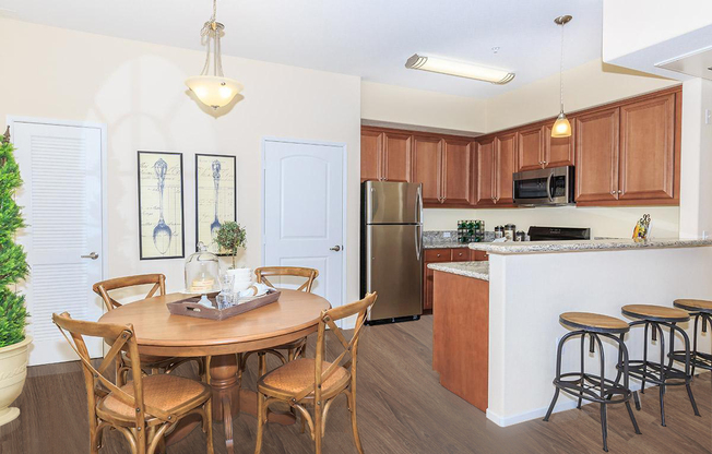A modern kitchen and dining area featuring a round wooden table with four chairs, a granite countertop, stainless steel appliances including a refrigerator and microwave, and warm wooden cabinets. Natural light fills the space, complemented by a potted plant and decorative wall art.