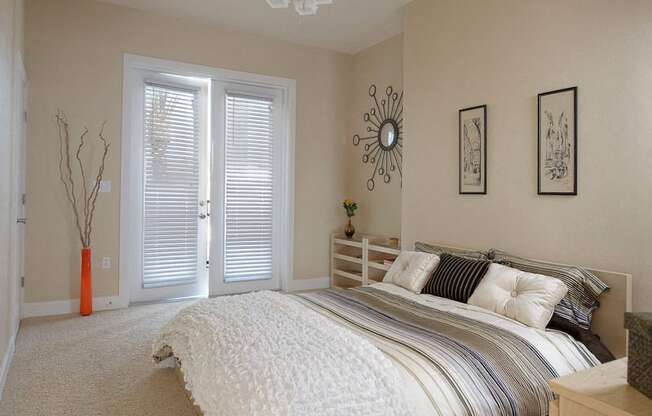 A staged bedroom with plush carpet flooring, ample natural light, and double doors to a private outdoor balcony.