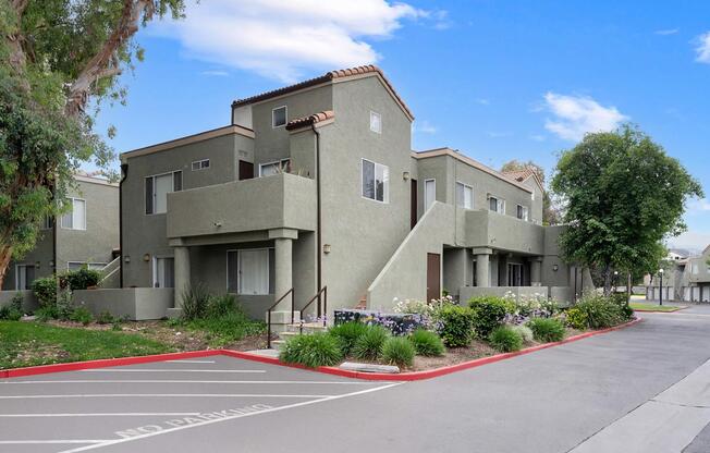 A multi-unit residential building with a light gray exterior. The structure features a tile roof, staircases leading to upper levels, and landscaped surroundings with shrubs and flowers. A "No Parking" sign is visible in the foreground, indicating designated parking areas. The sky is clear and blue.