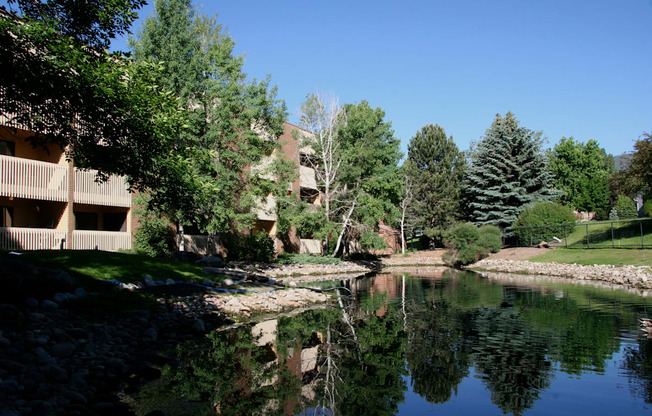 A peaceful landscaped pond here at Eagle Ridge reflecting surrounding trees and blue sky, with gently sloped grassy banks, natural stone edging, and mature evergreens alongside a brick apartment building with balconies, creating a serene outdoor setting that highlights the community’s greenery and scenic water feature.