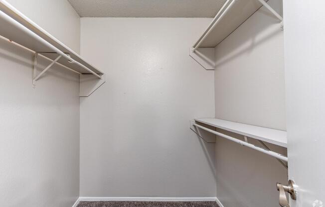 A clean, empty closet featuring white walls and a carpeted floor. It has two shelves mounted on the walls and a long shelf running along one side. The closet is well-lit and spacious, providing ample storage potential. The door is partially open, revealing the uncluttered interior.