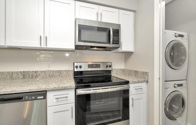 Modern kitchen featuring white cabinets, granite countertops, stainless steel appliances including an oven, microwave, and dishwasher. In the adjacent space, there is a stacked washer and dryer unit. The design is clean and contemporary, with a light color palette.