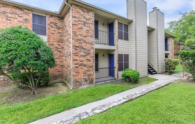 A two-story brick apartment building with a walkway in front, featuring balconies and green shrubs. The lawn is well-maintained with a concrete path leading to the entrance.