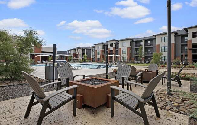 Modern 49 West Apartment courtyard featuring a fire pit surrounded by Adirondack chairs. A pool and lounge chairs are visible under a blue sky. Relaxed ambiance.