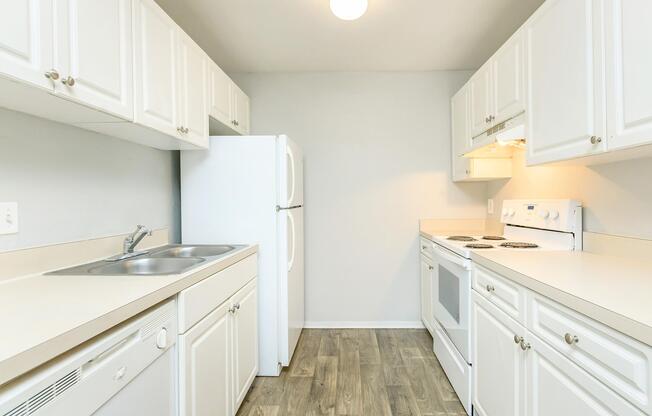 Bright and clean kitchen featuring white cabinetry, stainless steel sink, and a white refrigerator. The kitchen includes a stove and oven, with ample counter space and modern, light laminate flooring. The walls are painted in a light color, creating a spacious and inviting atmosphere.