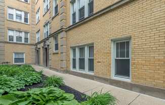 a brick apartment building with a sidewalk and plants in front of it