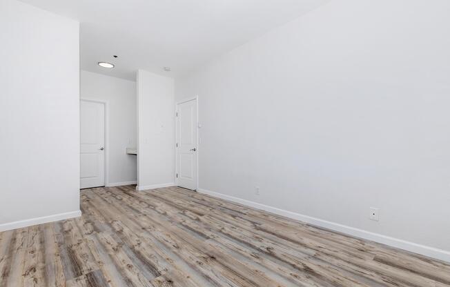 A spacious, empty room featuring light-colored walls and a wooden floor. A closed door is visible on the left leading to another space, while a second door on the right appears to lead to a bathroom or closet. The overall ambiance is bright and airy, with ample natural light.