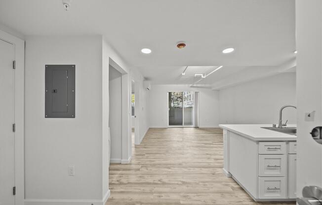 A bright, modern apartment interior showcasing an open layout. The view includes a kitchen island with white cabinetry, light hardwood flooring, and large windows allowing natural light. The space appears clean and inviting, with neutral-colored walls and minimalistic design elements.