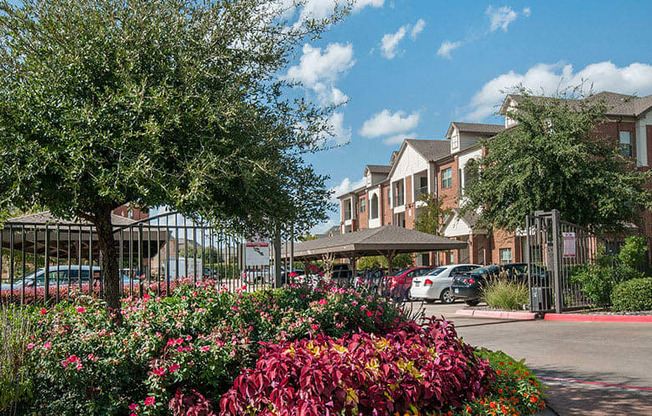 A community entrance and parking area here at Landing at Mansfield with gated access, covered carports, brick apartment buildings, and colorful landscaped flower beds and trees under a bright blue sky.