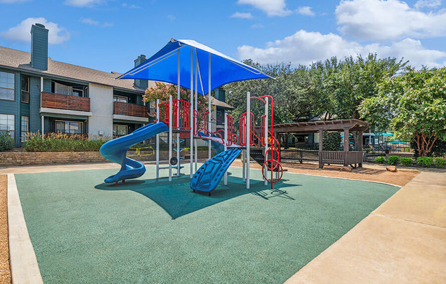 Community Playground with a Slide and Blue Canopy at Bridges at Deer Run Apartments in Dallas, TX.