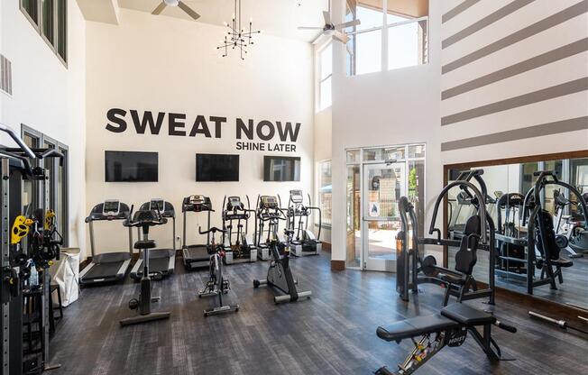 a gym with weights and cardio machines and a sweat now sign on the wall